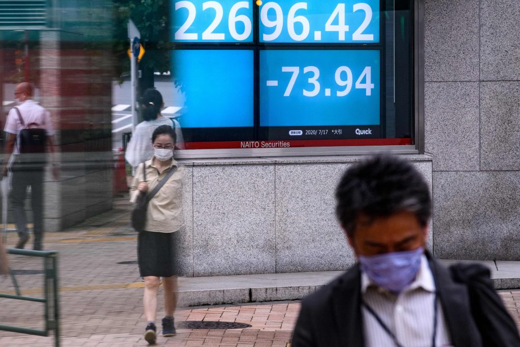 People wearing face masks walk across a road as an electric quotation board shows numbers of the Nikkei 225 index is seen in Tokyo, July 17, 2020. (File photo/AFP)