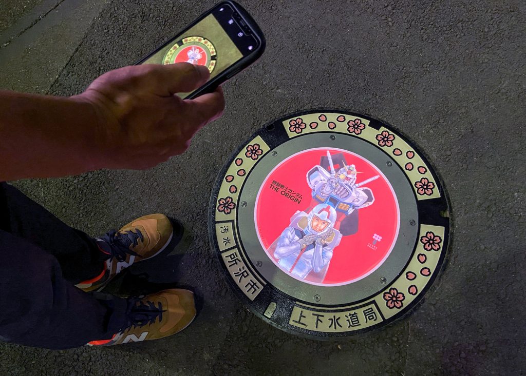 A passerby using a smartphone takes a photo of an illuminated manhole cover with designs of popular animation character Gundam, on the street in Tokorozawa, near Tokyo, Japan August 19, 2020. (Reuters)