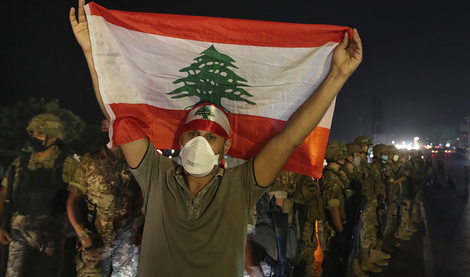An anti-government protester holds up a Lebanese flag as army soldiers stand guard during a demonstration against deteriorating economic conditions as politicians are deadlocked over forming a new government, in the town of Jal El-Dib, north of Beirut, Lebanon, Sunday, Sept. 27, 2020. (AP)