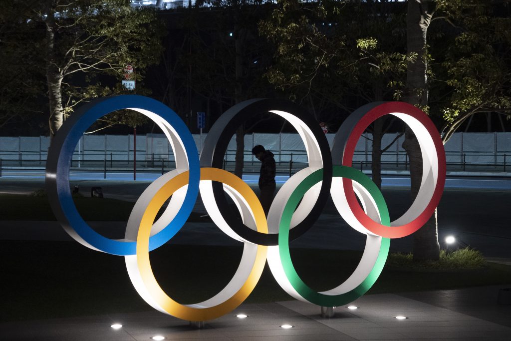 In this March 24, 2020, file photo, a man is seen through the Olympic rings in front of the New National Stadium in Tokyo. Tokyo Olympic organizers on Friday, Nov. 27, 2020 announced a series of 18 test events set to begin in March and run into May. (AP Photo/Jae C. Hong, File)