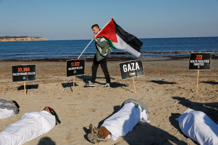 Activists wrapped in white sheets lie in the sand, while a person holds a Palestinian flag, during a symbolic protest action, as the conflict between Israel and the Palestinian Islamist group Hamas continues, at Ghadira Bay in Mellieha, Malta, February 3, 2024. (Reuters)