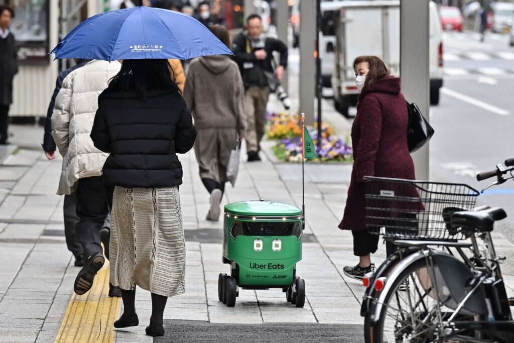Pedestrians walk alongside an unmanned robot (C) as it navigates down a street during a demonstration of a robot delivery service by Uber Eats Japan, Mitsubishi Electric and robot developer Cartken in downtown Tokyo on March 5, 2024. (Photo by Richard A. Brooks / AFP)