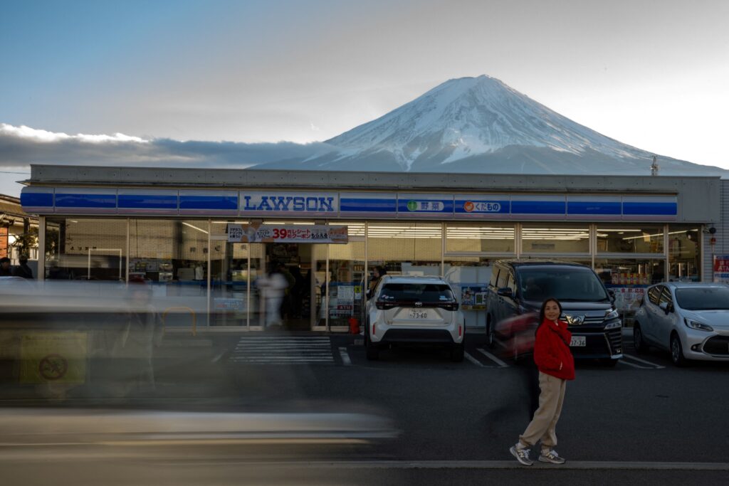 This photo taken on January 1, 2024 shows a tourist posing in front of a convenience store with Mount Fuji in the background, in the town of Fujikawaguchiko, Yamanashi prefecture. A huge black barrier to block Mount Fuji from view will be installed in a popular photo spot by Japanese authorities exasperated by crowds of badly behaved foreign tourists, it was reported on April 26, 2024. (Photo by Philip FONG / AFP)