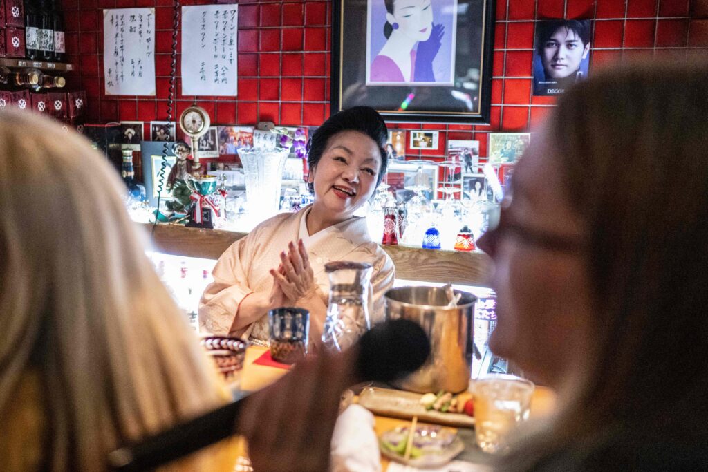 Behind the bar, dressed in a smart pale kimono with her hair in a traditional up-do, is 