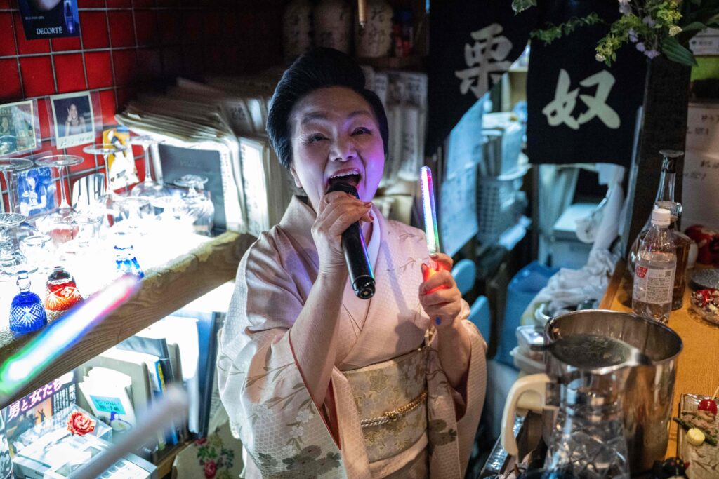 Behind the bar, dressed in a smart pale kimono with her hair in a traditional up-do, is 