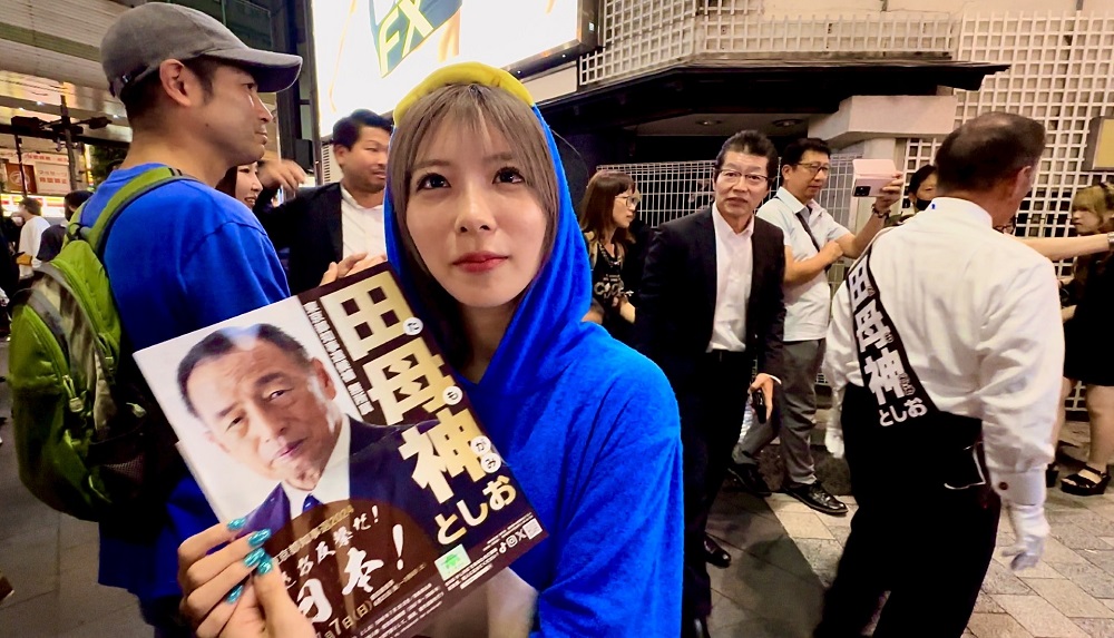 Popular TikToker Yukinan holds a poster of Tokyo governor candidate, TAMOGAMI Toshio who appears in the background after he delivered a campaign speech in Tokyo's Roppongi district. (ANJ)