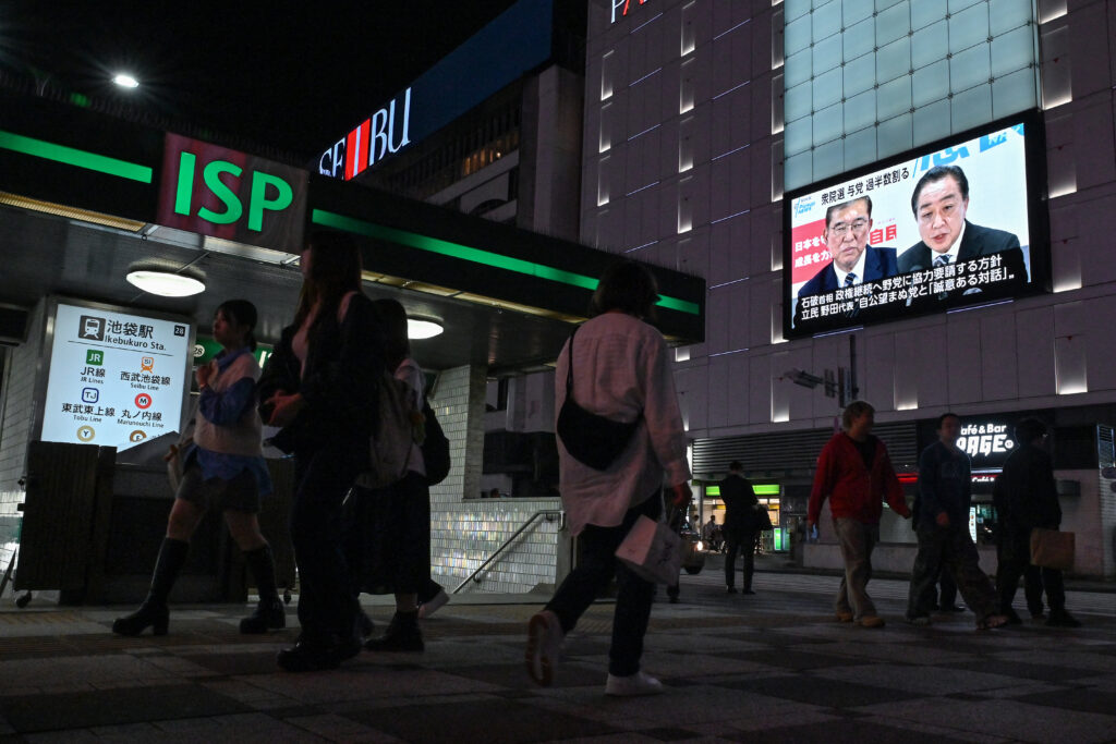 People walk outside Ikebukuro train station as images of Japan's Prime Minister Shigeru Ishiba (top L) and former premier and head of the opposition Constitutional Democratic Party (CDP), Yoshihiko Noda (top R) are broadcast during a news update on a large television screen one day after the general election in Tokyo on October 28, 2024. (AFP)