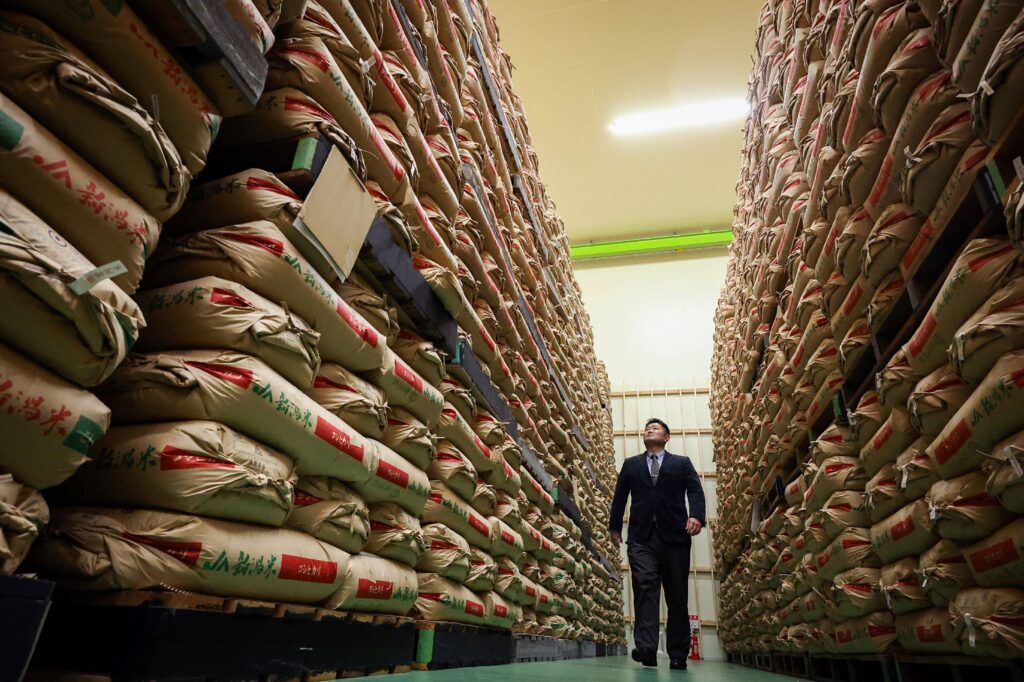 This file photo taken on February 18, 2025 shows a man walking past sacks of government stockpiled rice piled up in a warehouse in Saitama prefecture, north of Tokyo. Japan's new farm minister said on May 26, 2025 the country's embattled government would release reserve rice directly to large retailers in an attempt to bring down prices for consumers after the recent spike. (Photo by JIJI Press / AFP) 