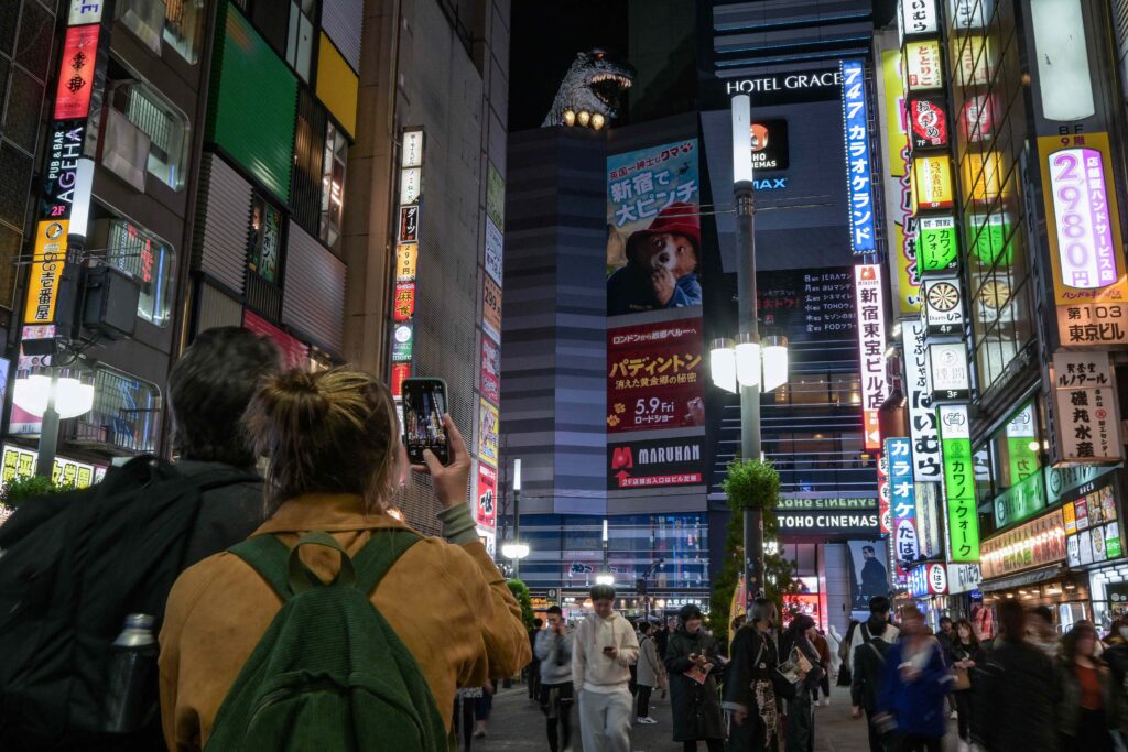 This photo taken on April 8, 2025 shows a tourist (L) taking a photo of a figure of classic film monster Godzilla (top C) pictured at the top of a building in the red-light entertainment area of Kabukicho in the Shinjuku district of Tokyo. (AFP)