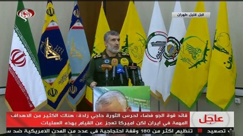 国営テレビ番組にて、公式のイラン国旗と並んで掲げられるさまざまな代理組織の旗の前に立つアミール・アリ・ハジザデ司令官（画像）