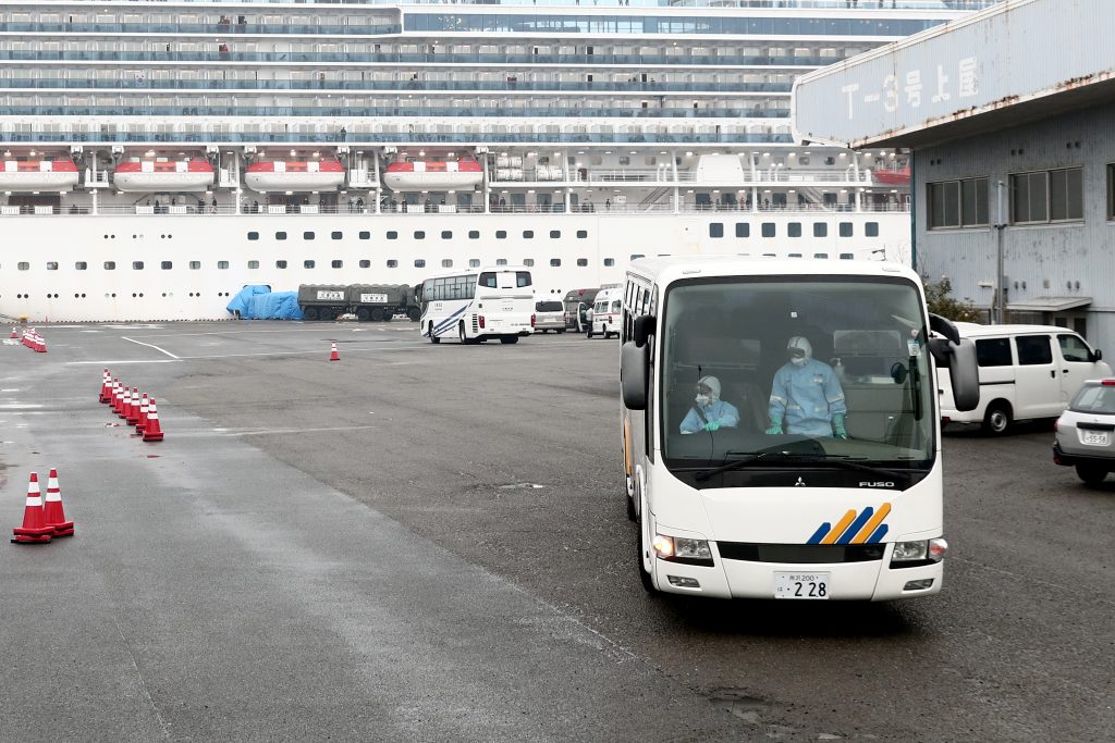 防護服を着た運転手(左)が乗ったバスがクルーズ船ダイヤモンド・プリンセス号が停泊する横浜港の大黒ふ頭客船ターミナルの埠頭を出発する場面。同船では乗っている約3,600人が新型のCOVID-19コロナウイルスのおそれにより隔離されている。(AFP)