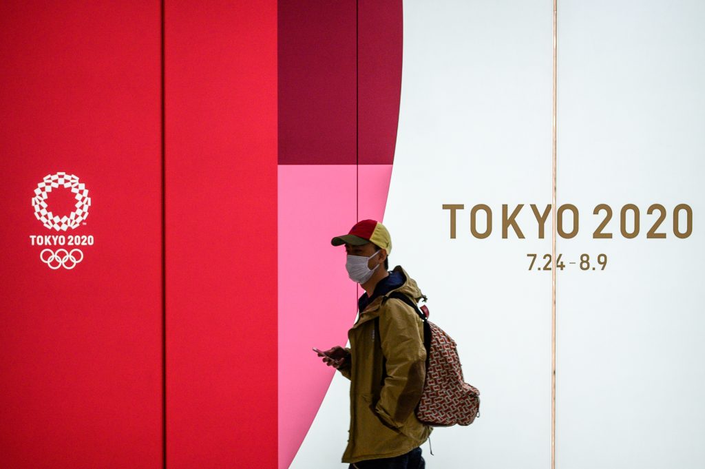 2020年3月11日、東京にて。2020年夏季五輪の公式の広告を背景に、地下鉄の駅の通路をマスクを着用して歩く男性。（AFP）