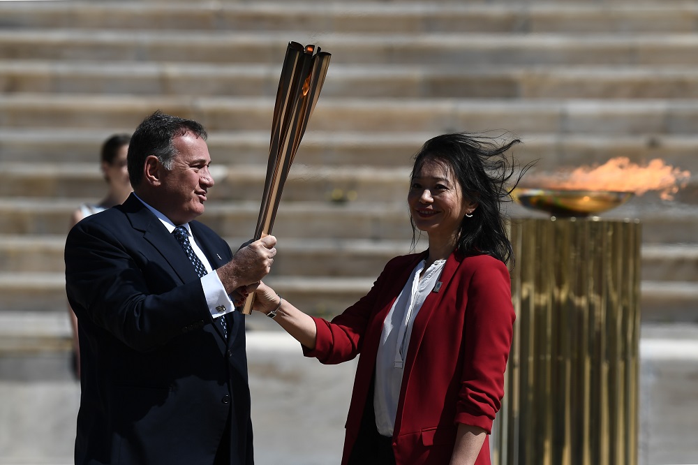 2020年3月19日にアテネで開催された、2020年東京夏季オリンピックの聖火引継式で、ギリシャのスポーツ大臣でありHOC会長であるスピロス・カプラロス（左）が、日本の元水泳選手の井本直歩子にオリンピックの聖火を手渡す。（AFP）