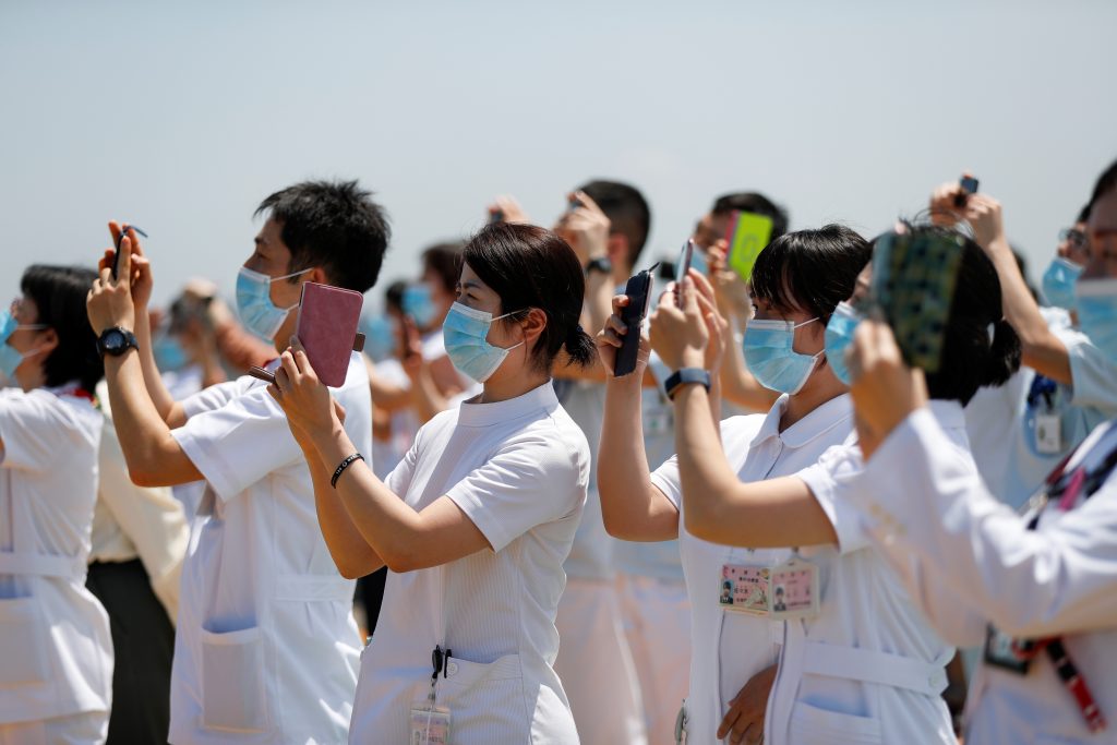 2020年5月29日、日本の東京で日本の航空自衛隊のアクロバットチーム「ブルーインパルス 」が新型コロナウイルス感染症（COVID-19）との闘いの最前線にいる医療従事者に敬意を示すために自衛隊中央病院の上空を飛ぶのを見る、携帯電話を持った医療従事者たち（ロイター）。