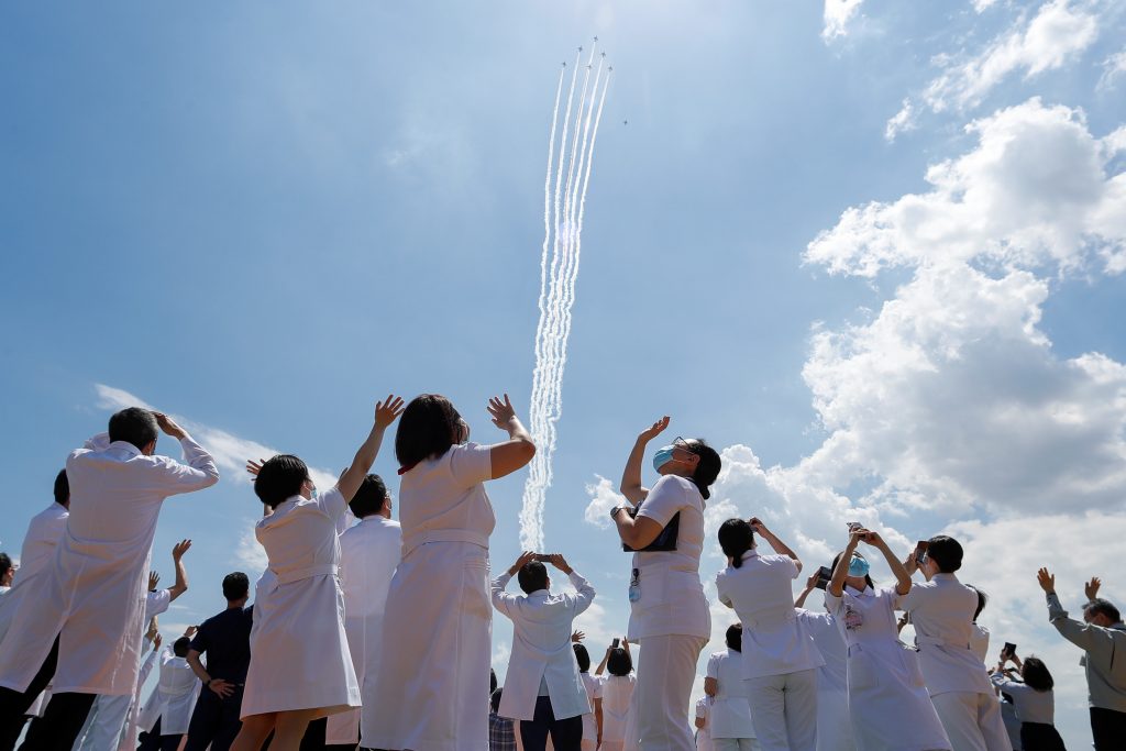 2020年5月29日、日本の東京で日本の航空自衛隊のアクロバットチーム「ブルーインパルス 」が新型コロナウイルス感染症（COVID-19）との闘いの最前線にいる医療従事者に敬意を示すために自衛隊中央病院の上空を飛ぶのを見て反応する医療従事者たち（ロイター）。