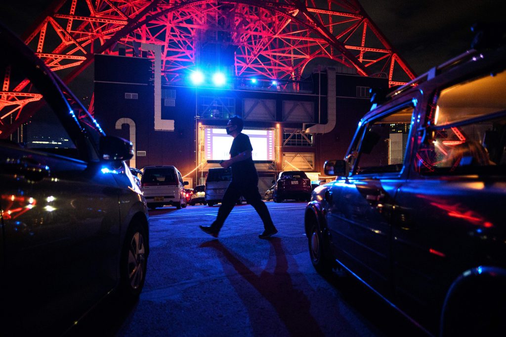 東京で行われたドライブインシアター・イベント中、駐車場を歩くマスク姿の男性。このイベントはCOVID-19コロナウィルス・パンデミックにより社会的制約が課せられる中の2020年6月20日、東京タワーの真下で開催された。（AFP）