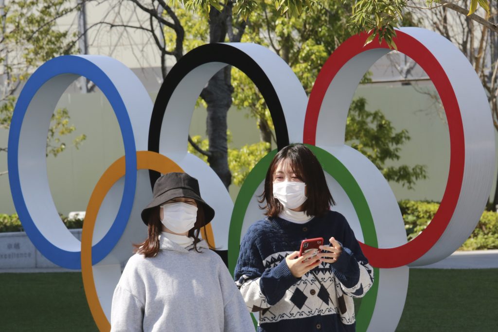 2021年3月17日、東京で五輪マークの前を歩く人々の写真。東京オリンピックの開幕まであと4ヶ月を切り、1万人のランナーによる聖火リレーが日本を縦断し始めた。主催者はリスクを軽減していると言うが、多くの医学専門家は慎重な姿勢を崩していない。(AP通信写真／サシハラ・コウジ)