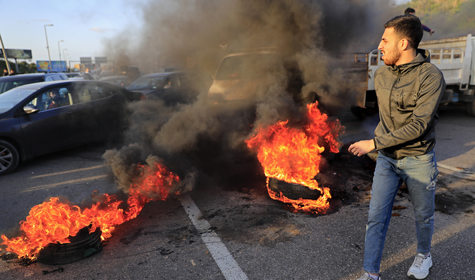 2021年3月2日、レバノンのベイルートで、人々が車の中で身動きが取れなくなる中、ベイルートの国際空港へと続く幹線道路を封鎖するために火がつけられたタイヤの前を通り過ぎる抗議活動家。（AP通信）