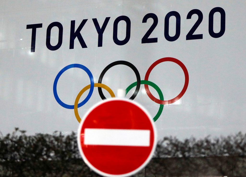 新型コロナウイルス感染症(COVID-19)の感染拡大により、2021年に延期された東京2020オリンピックのロゴが、日本の東京都庁の交通標識越しに見える=2021年1月22日(ロイター通信/カトウ・イッセイ)