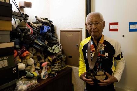 2021年8月2日に撮られたこの写真は、千葉県稲毛区の自宅にて積み上げられた靴の山の隣で撮影のためのポーズをとる世界最高齢のアイアンマン競技者である稲田弘さんを収めたもの。（AFP）