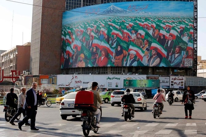 2022年10月25日、イランの首都テヘランの中心部のヴァリ・アスル広場で横断歩道を渡る人々。巨大な広告用掲示板には、さまざまな職業や地位の男女がイランの国旗とファーティマ・アル・ザハラ（預言者ムハンマドの娘）、イマーム・フサイン（ムハンマドの孫）、預言された救世主マフディーの名が書かれた緑色の旗を手に行進する壁画が掲げられている。（Photo by AFP）