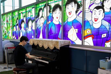 この資料写真は2020年7月4日に撮影されたもので、新型コロナウイルスの影響で延期されていたJリーグの試合が再開された日に、埼玉県の浦和美園駅にて、漫画『キャプテン翼』のキャラクターを描いたステンドグラス作品の横でピアノを演奏する旅客者が写っている。（AFP）