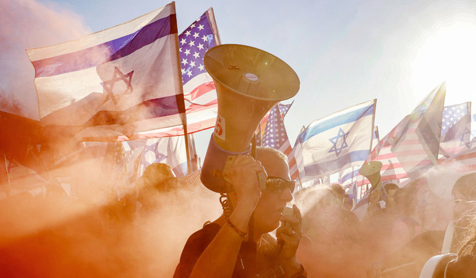 2023年7月11日、テルアビブの米在外公館のテルアビブ支公館前にて、イスラエル政府の司法改革法案に反対する抗議デモの最中、ある抗議デモ参加者が、発煙筒の煙の中をイスラエルと米国の国旗を掲げた人々の前を通りながら、メガホンで訴えているところ。（AFP）