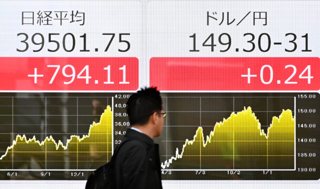 東京証券取引所の株価（左）と日本円対ドルレート（右）を示す電光掲示板の前を通り過ぎる男性（2024年3月18日、東京の路上で）。(AFP＝時事）