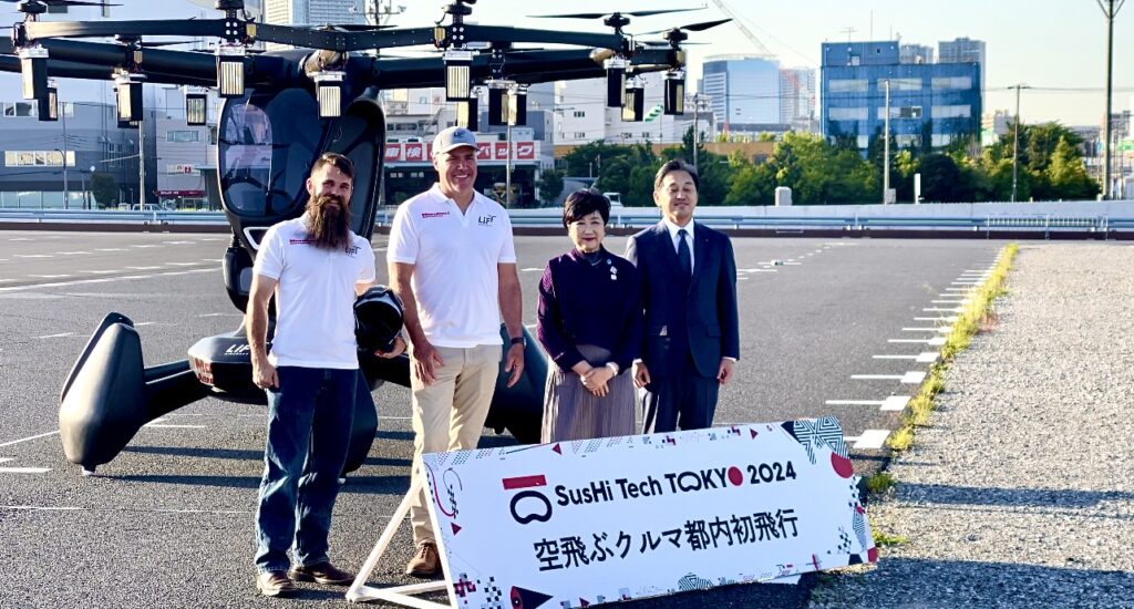 フライング・カーの機長(右端)、フライトアシスタント、東京都職員とともに報道陣の取材に応じる小池百合子東京都知事(ANJ)。